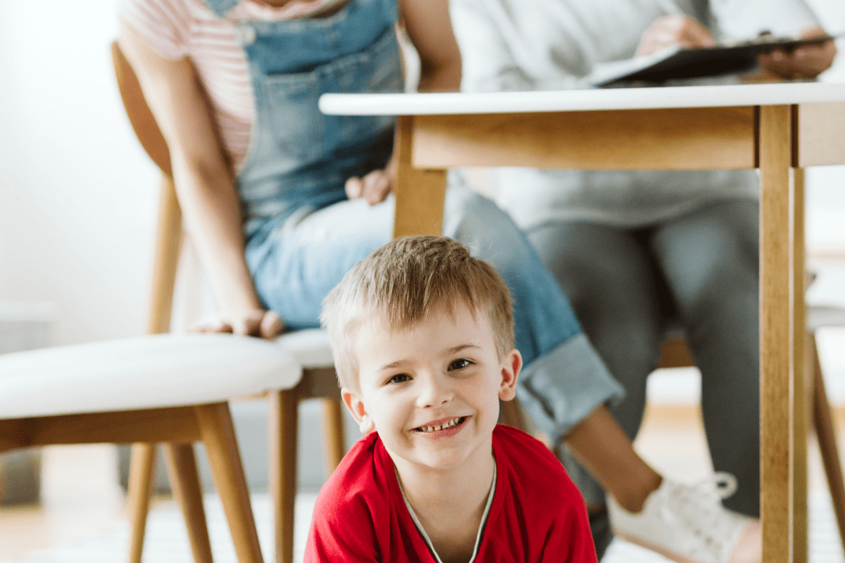 Young child smiling on the floor while adults talk at a table
