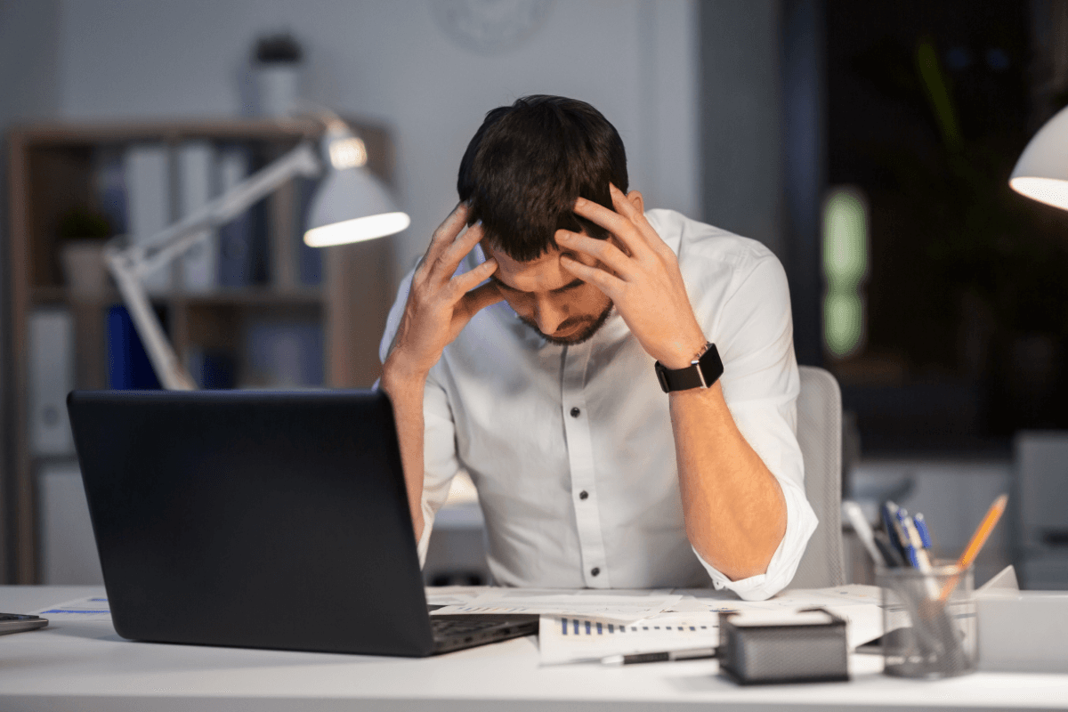 Man at a laptop holding his head over a desk of papers
